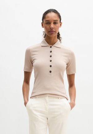 Femme aux cheveux bouclés portant un polo côtelé beige à manches courtes avec des boutons et un pantalon blanc, debout les mains dans les poches sur un fond blanc.