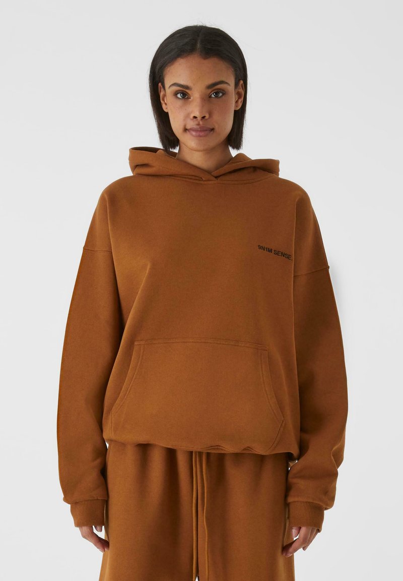 Brun oversized hettegenser med en frontlomme, justerbar hette og ribbestrikkede mansjetter. Har minimal svart tekst på brystet. Laget av myk bomullsblanding.
