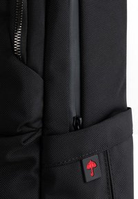 Czarny plecak z teksturowanej tkaniny, z zamykaną na zamek kieszenią oraz czerwoną parasolką na naszywce. Minimalistyczny design z praktycznymi detalami.