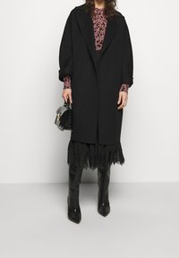 Abrigo de lana negro con solapas oversized, combinado con una blusa floral rosa, falda de encaje en capas y elegantes botas negras hasta la rodilla. Bolso compacto negro.
