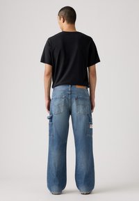 Jeans de mezclilla azul holgados con cintura alta, que cuentan con dos bolsillos traseros y dos bolsillos laterales, combinados con una camiseta negra de manga corta.