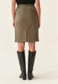 Jupe kaki arrivée au genou avec fente à l'arrière, dotée de deux poches arrière avec zippers, associée à des bottes en cuir noires arrivant au genou.