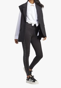 Černé legíny s texturovaným potiskem po stranách, spárované s bílým tričkem uvázaným v pase a bezrukávovým černým blejzrem. Černé tenisky.