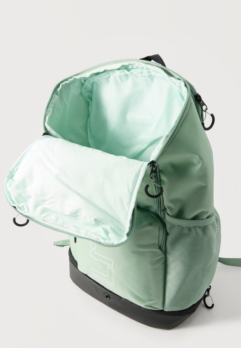 Mochila verde claro con compartimento principal abierto que muestra el forro interior claro, bolsillo lateral de malla y base negra sobre fondo blanco.
