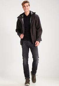 Black waterproof jacket with a hood, layered over a black shirt, paired with dark jeans and olive green sneakers, featuring a zip front.