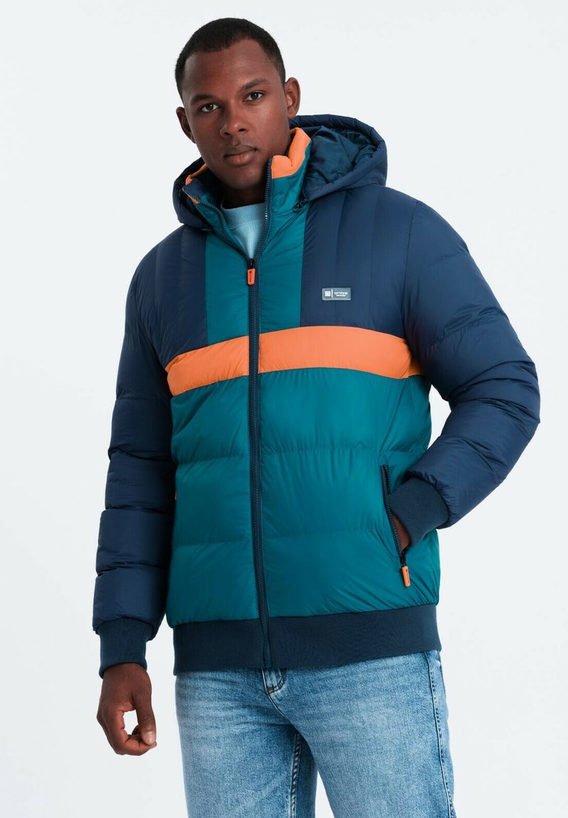 En pufferjacka i teal och marinblå med ett orange rand. Har en huva, dragkedjestängning och ribbade ärmslut. Mjuk, quiltad textur.