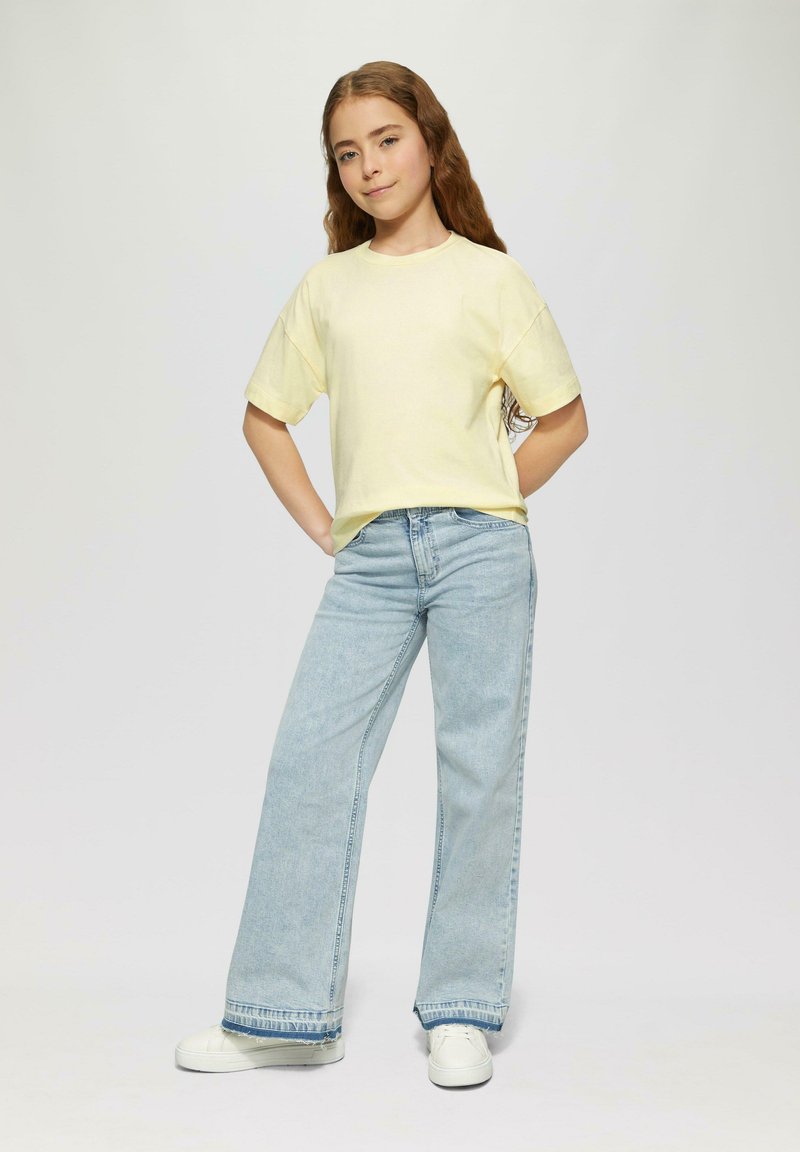 Helles gelbes Kurzarm-T-Shirt, lockere Passform. Hellblaue weit geschnittene Jeans mit fransigem Saum. Weiße Sneakers. Lässiges, entspanntes Outfit.