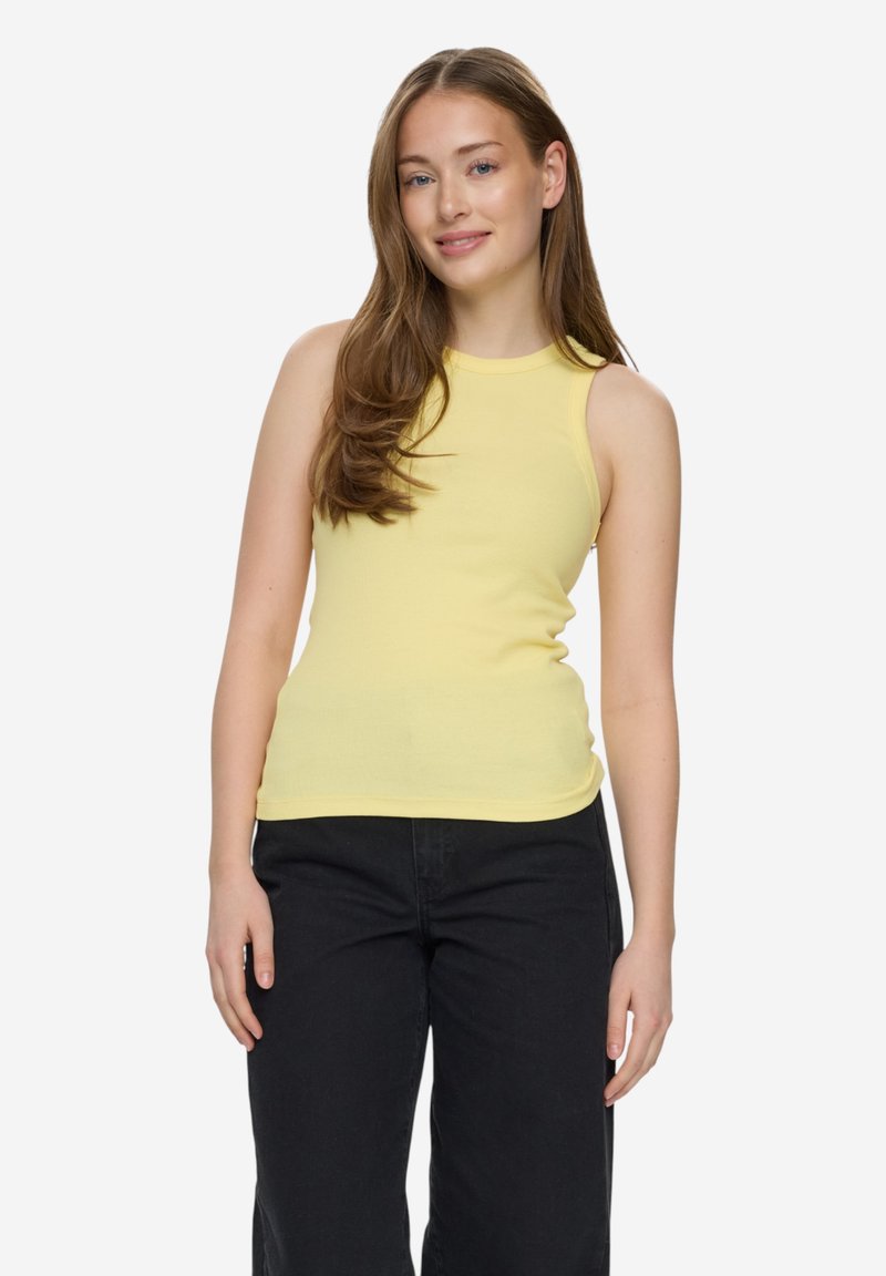 Jeune femme aux longs cheveux bruns, vêtue d'un haut jaune sans manches et d'un pantalon noir, debout et souriante devant un fond blanc.