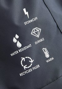 Un tissu gris foncé présente des icônes blanches pour le manchon tempête, la résistance à l'eau, la durabilité, le rembourrage recyclé et la chaleur, accompagnées de descriptions textuelles simples.