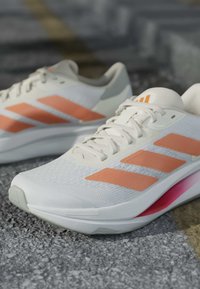Chaussures de sport blanches avec des rayures orange et un accent rose sur la semelle, placées sur une surface grise texturée.