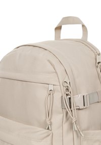 Zaino in tessuto beige con forma arrotondata, tasca frontale, corde di regolazione e maniglia superiore. Cerniera e accessori dello stesso colore chiaro.