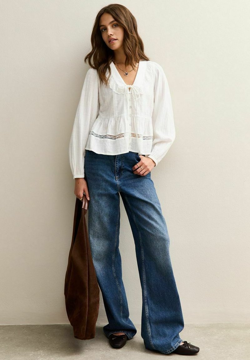 Vrouw in een losse witte blouse en wijde blauwe jeans, met een grote bruine tas, staand tegen een effen muur.