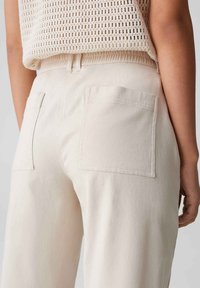 Pantalon en coton beige avec une coupe décontractée, doté de poches arrière à patch et d'une texture lisse. Porté avec un haut en tricot ample.