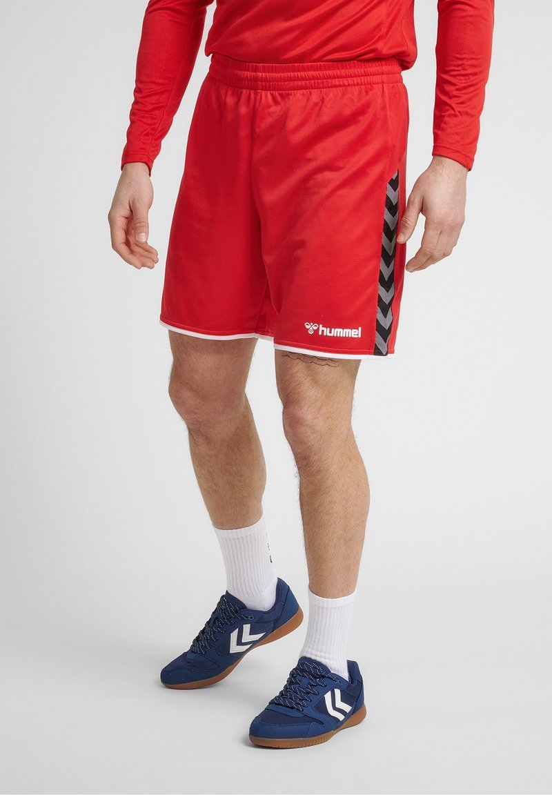Shorts de sport rouges avec un motif en chevron noir sur le côté, ornés d'un liseré blanc et d'une taille élastique. Portés avec des baskets bleues.