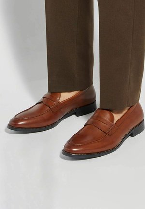 Bruine leren loafers met een gladde textuur, voorzien van een gestikt ontwerp aan de voorkant en een lage zwarte hak. Gecombineerd met bruine broek.