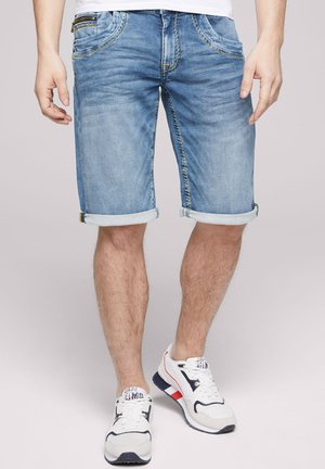 Shorts en denim bleu clair avec ourlet retroussé, présentant un léger délavage, des coutures jaunes et des poches avant zippées. Associés à des baskets blanches.