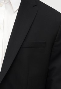 Κοντινό πλάνο ενός ατόμου που φοράει ένα μαύρο σακάκι πάνω από ένα λευκό πουκάμισο με γιακά, εμφανίζοντας το πέτο και την τσέπη στον θώρακα του σακακιού.