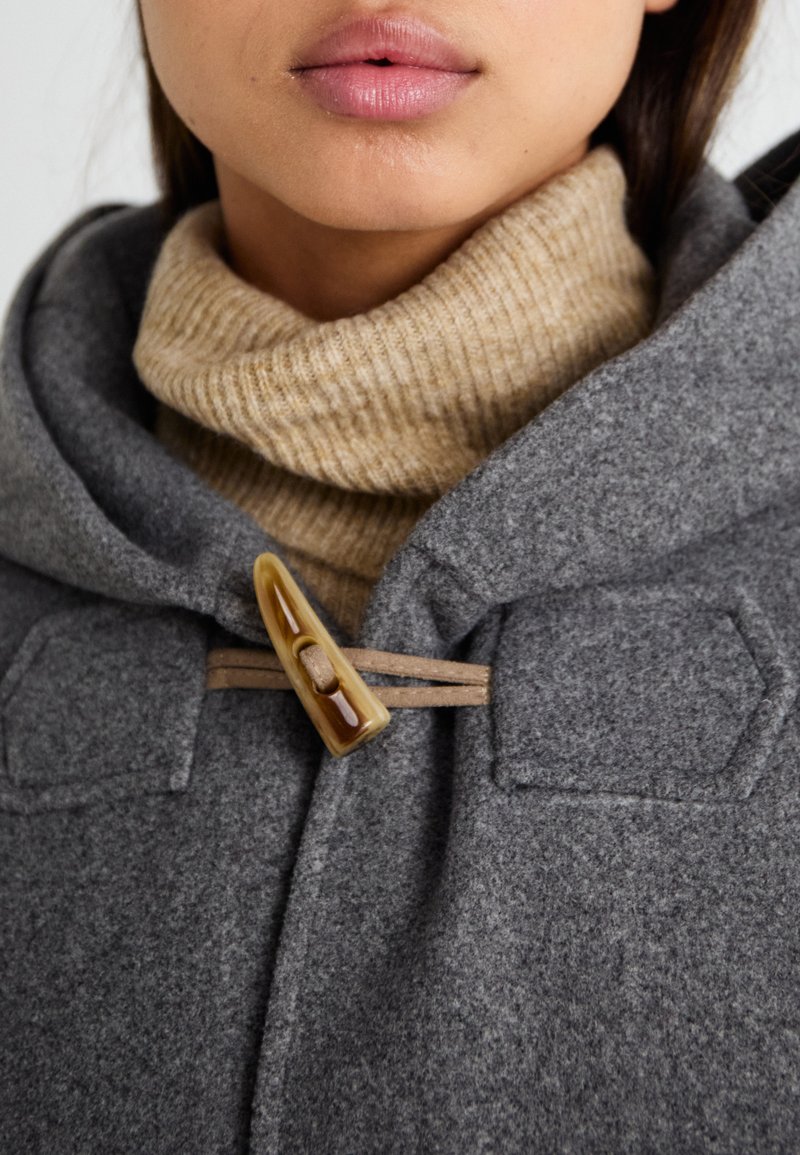 Szary wełniany płaszcz z kapturem, z brązowym zapięciem na zaczepy i beżowym prążkowanym golfem pod spodem. Teksturalna tkanina i heksagonalne kieszenie na łatki.