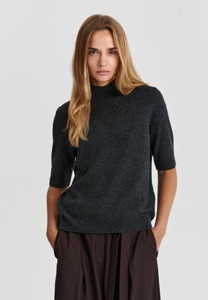 Femme aux longs cheveux blonds portant un pull gris foncé à manches courtes et un pantalon large foncé, debout les mains dans les poches devant un fond uni.