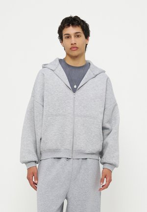 Giovane adulto che indossa una felpa con cappuccio grigia chiara con zip e pantaloni della tuta abbinati, con una maglietta grigio scuro e una collana con ciondolo rotondo.