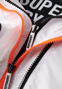 Biała kurtka z pomarańczowymi i czarnymi zamkami błyskawicznymi, z textured wewn�ętrzną podszewką i logo "SUPERDRY" na uchwycie zamka.