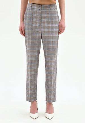 Pantalon à carreaux beige et bleu, coupe ajustée, longueur cheville, porté avec des escarpins blancs à talons pointus sur une personne debout.