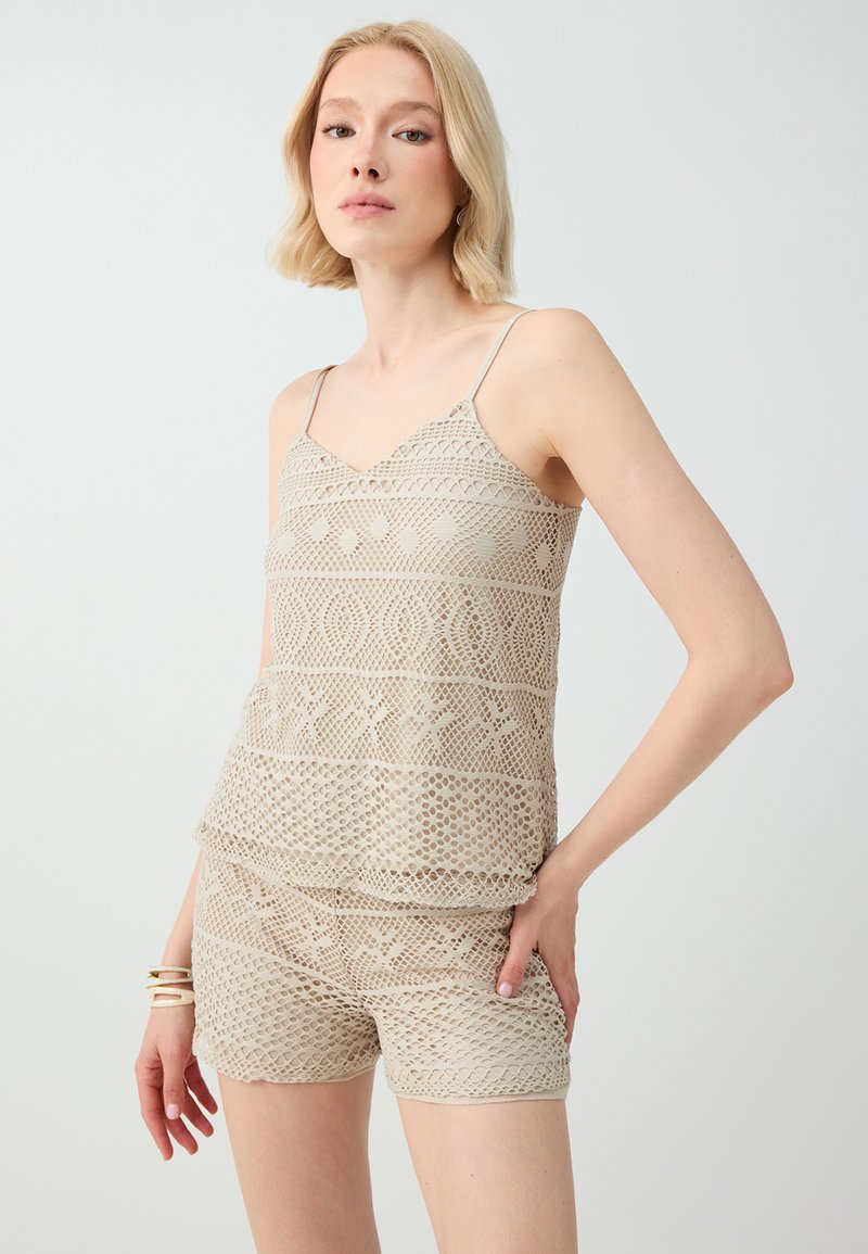 Femme blonde portant un caraco en crochet beige et un short assorti avec des motifs géométriques, posant devant un fond clair uni.