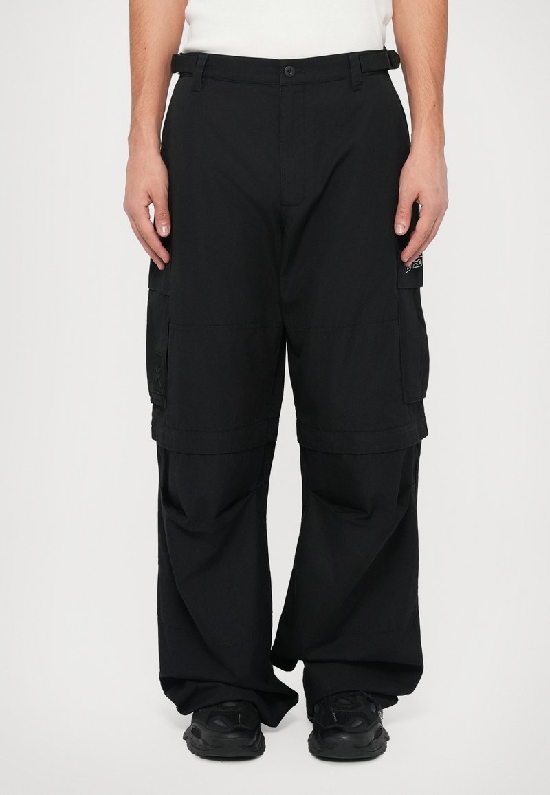 Schwarze Cargo-Hosen mit lockerer Passform, mehreren seitlichen Taschen und glatter Textur, kombiniert mit schwarzem Schuhwerk.