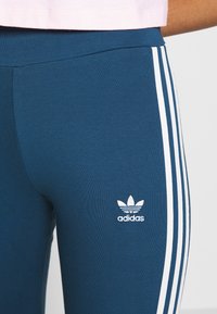 Modré legíny s bílými postranními pruhy. Na levém stehně je vyšitý bílý logo Adidas. Hladká, elastická tkanina.