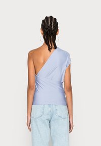 Vrouw met gevlochten haar, die grote oorbellen draagt, een licht paarse top met één schouder en lichtblauwe spijkerbroek, met haar rug naar de camera op een witte achtergrond.
