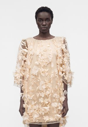 Γυναίκα με κοντά σγουρά μαλλιά φορώντας μπεζ διαφανές φόρεμα διακοσμημένο με 3D λουλουδένια εφαρμογές και κεντήματα, στέκεται μπροστά σε ένα απλό φόντο.