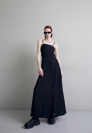 Czarna długa sukienka na cienkich ramiączkach, dopasowany gorset i zwiewna spódnica. Modelka nosi masywne czarne platformy i małe czarne okulary przeciwsłoneczne.