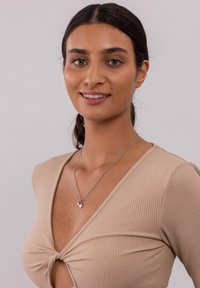 Silberne Halskette mit einem herzförmigen Anhänger, getragen mit einem beigefarbenen, gerippten Oberteil mit einer Drehdetails. Das Modell hat einen neutralen Hintergrund.