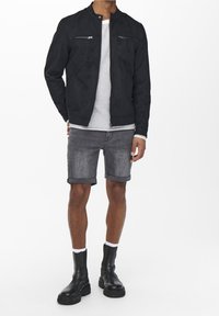 Giacca in suede nero con chiusura a zip e due tasche sul petto, abbinata a pantaloni corti in denim grigi e stivaletti neri con suola spessa.