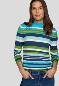 Frau mit braunen Haaren, die einen gestrickten Pullover in Blau, Grün und Weiß trägt, die Hände in den Taschen, vor einem einfarbigen hellgrauen Hintergrund.