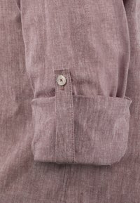 Camisa de manga larga en una tela de color malva claro con una textura suave. Presenta un detalle de botón en el puño y costuras visibles a lo largo de las costuras.