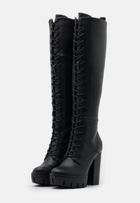 Bottes plates-formes noires montant jusqu'au genou en cuir lisse, avec un design à lacets sur le devant et des semelles épaisses et texturées.