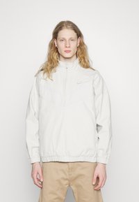 Lysegrå zip-up Nike jakke med høj hals, elastiske manchetter og en horisontal sømdetalje. Blødt stof med diskret logo-accs.