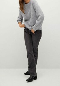 Femme portant un pull en tricot gris, un jean noir à jambes droites et des bottines à talons noires, se tenant devant un fond clair et uni.