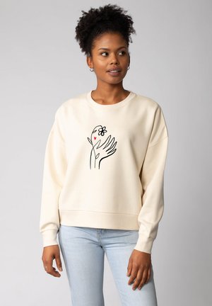 Crème sweatshirt gemaakt van zachte stof, met een zwarte lijntekening van een hand met een bloem en een hart op de voorkant. Ontwerp met een relaxte pasvorm.