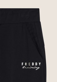 Pantalones deportivos negros de tejido suave con un bolsillo lateral y un logotipo plateado que dice "FREDDY training" en fuentes contrastantes.