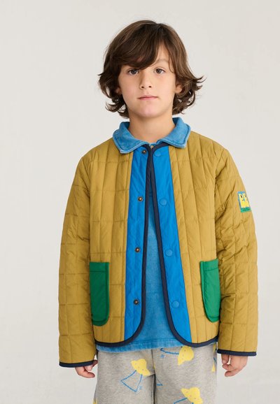 Enfant aux cheveux bruns portant une veste matelassée jaune avec des poches vertes et des bordures bleues sur une chemise bleue et un pantalon gris avec des motifs jaunes.