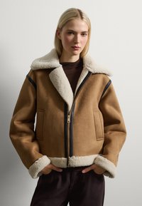 Chaqueta de ante marrón con cuello y puños de piel sintética en crema, cierre frontal con cremallera y bolsillos laterales. Presenta detalles en negro con cremallera.