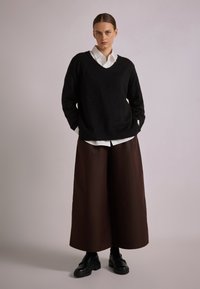 Black knit sweater with v-neck, worn over a white button-up shirt. Paired with a long, brown skirt and black chunky shoes.