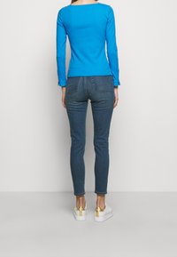 Blå långärmad topp med fyrkantig halsringning, kombinerad med mörka tight jeans. Den avslappnade outfiten kompletteras av vita sneakers med gulddetaljer.