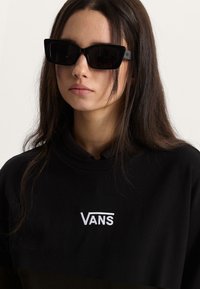 Lunettes de soleil noires à monture œil de chat avec une finition mate, présentant un design audacieux et la marque Vans sur la branche, portées avec un sweat-shirt noir.