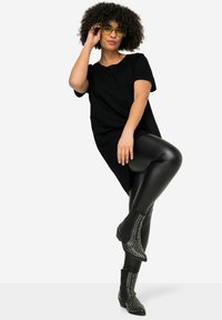 T-shirt noir oversize, leggings noirs brillants et bottines noires à clous. Le mannequin pose avec des cheveux bouclés et des lunettes de soleil jaunes.