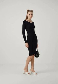 Czarna sukienka bodycon z długim rękawem i okrągłym dekoltem. Gładka tekstura, do kolan. Połączona z białymi sandałami na obcasie z odkrytą palcą i czarną torebką.