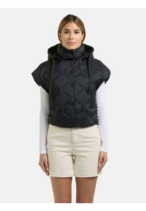 Kvinde iført en sort quiltet, ærmeløs hættetrøje over en hvid langærmet top og beige shorts, stående foran en ensfarvet baggrund.