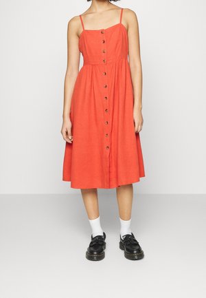 Femme portant une robe midi orange sans manches avec des boutons sur le devant, chaussettes blanches et chaussures noires à lacets sur un fond uni.